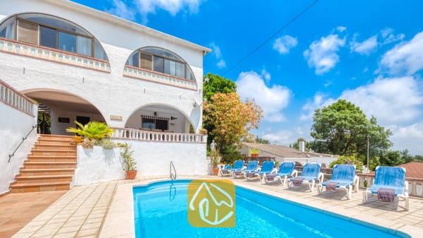 Vakantievilla's Costa Brava Spanje - Villa Santa Maria - Buitenzwembad en witte buitenkant van Villa Santa Maria in Spanje, met blauwe ligstoelen, boogvormige ramen en tegelいません en betegelde trappen onder een blauwe lucht met wolken.