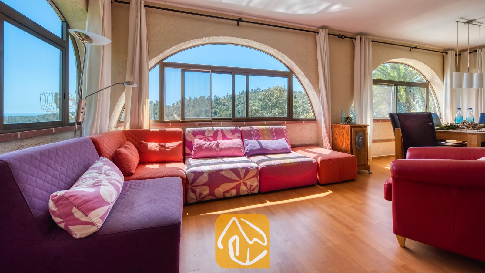 Vakantievilla's Costa Brava Spanje - Villa Santa Maria - Lichte woonkamer in Villa Santa Maria met een grote hoekbank met paarse en roze bloemenprints, houten vloeren en boogvensters die uitkijken op groene heuvels.