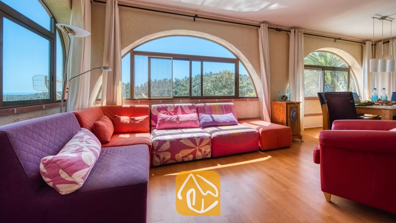 Vakantievilla's Costa Brava Spanje - Villa Santa Maria - Lichte woonkamer in Villa Santa Maria met een grote hoekbank met paarse en roze bloemenprints, houten vloeren en boogvensters die uitkijken op groene heuvels.