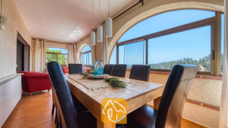 Vakantievilla's Costa Brava Spanje - Villa Santa Maria - Eethoek in Villa Santa Maria met een houten tafel, donkerlederen stoelen en grote boogvormige ramen met uitzicht op bomen en heuvels.