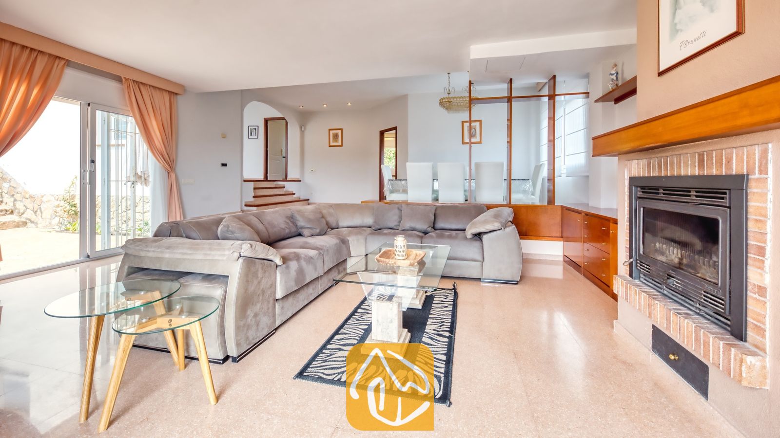 Vakantievilla's Costa Brava Spanje - Villa Valentina - Ruime woonkamer in Villa Valentina met een grote grijze hoekbank, een stenen open haard en glazen salontafels op een bedrukt vloerkleed.