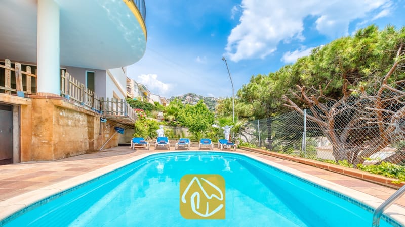 Vakantievilla's Costa Brava Spanje - Villa Valentina - Buitenzwembad bij Villa Valentina met blauw water, omringd door tegelbestrating, vijf ligstoelen en dennenbomen onder een blauwe lucht.