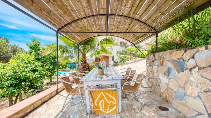 Vakantievilla's Costa Brava Spanje - Villa Valentina - Overdekte buiten-eethoek bij Villa Valentina met een lange witte houten tafel en geweven rieten stoelen onder een gebogen houten pergola van latten.