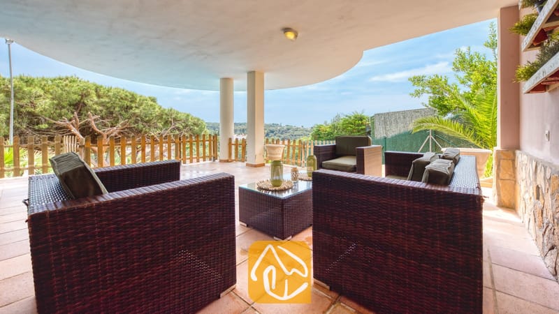 Vakantievilla's Costa Brava Spanje - Villa Valentina - Overdekt terras bij Villa Valentina met bruine rieten fauteuils, een salontafel met glazen blad en tegelvloeren die uitkijken op groene bomen en een blauwe lucht.