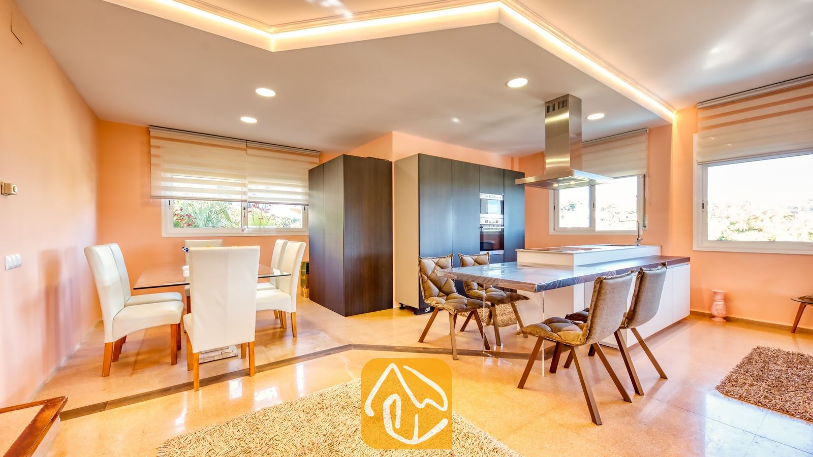 Vakantievilla's Costa Brava Spanje - Villa Valentina - Moderne keuken en eethoek in Villa Valentina met donkere kasten, een glazen eettafel met witte stoelen en een kookeiland met gestoffeerde bruine stoelen.