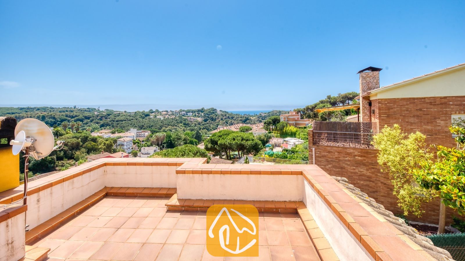 Vakantievilla's Costa Brava Spanje - Villa Valentina - Terracotta betegeling, witte muren met bruine dakranden en een stenen schoorsteen vanaf het terras van Villa Valentina in Spanje, met uitzicht op groene heuvels en een stad in de verte.