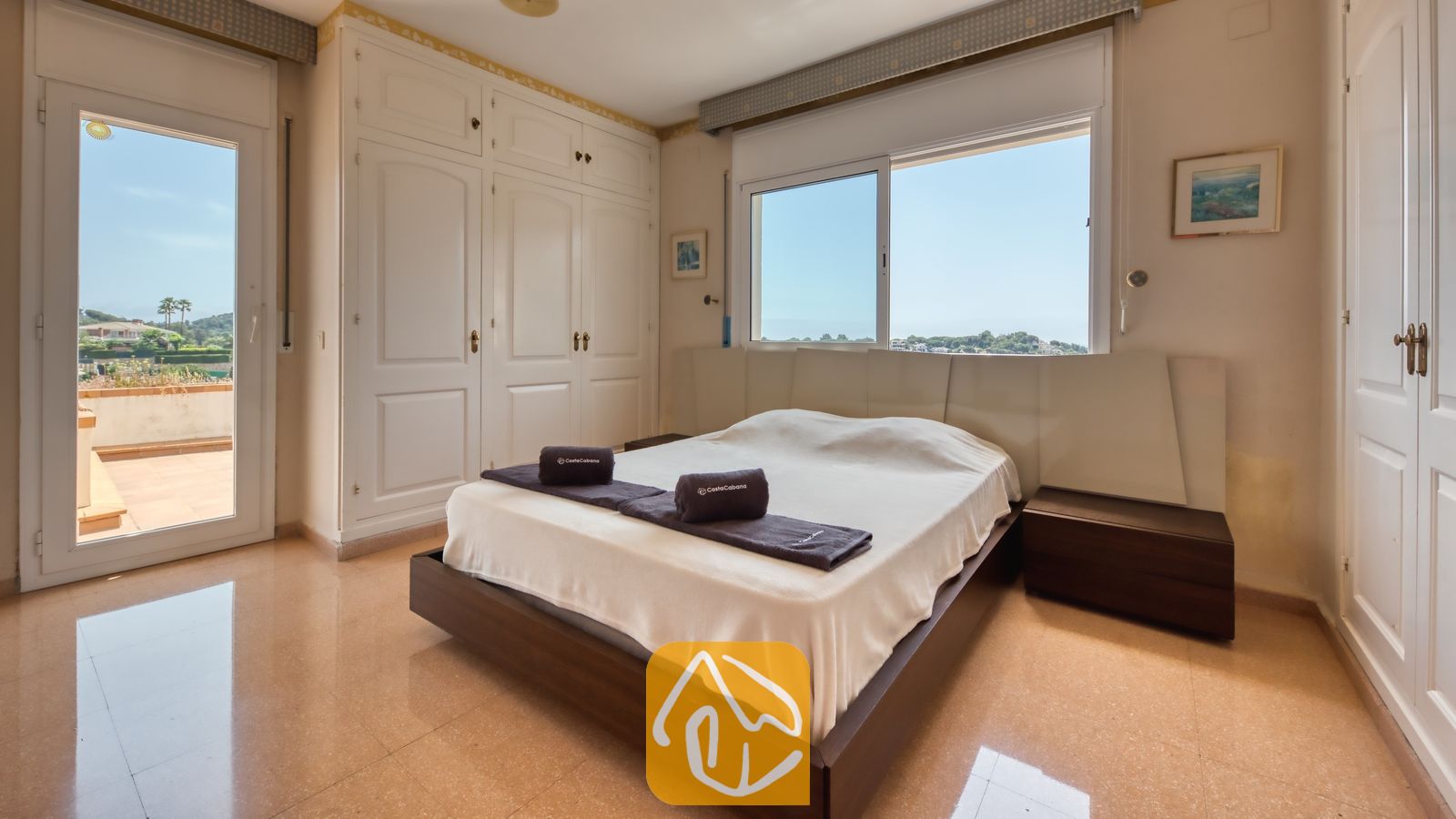 Vakantievilla's Costa Brava Spanje - Villa Valentina - Slaapkamer met tweepersoonsbed, donkerhouten ombouw, witte kledingkasten, raam met uitzicht op bomen en de lucht, en een balkondeur.