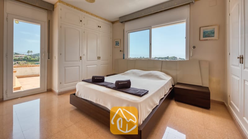 Villas de vacances Costa Brava Espagne - Villa Valentina - Chambre avec lit double, cadre en bois sombre, armoires blanches, fenêtre offrant une vue sur les arbres et le ciel, et porte-fenêtre donnant sur le balcon.