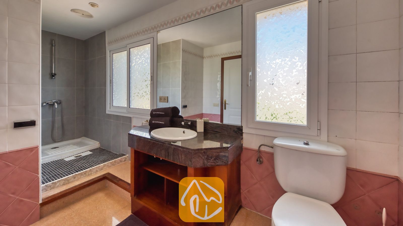 Vakantievilla's Costa Brava Spanje - Villa Valentina - Badkamer met een donker granieten wastafelmeubel, een witte wastafel, een houten onderkast, een betegelde inloopdouche en matglas raam.