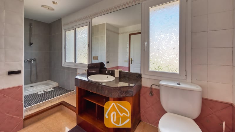 Villas de vacances Costa Brava Espagne - Villa Valentina - Salle de bains dotée d'un meuble-vasque en granit sombre, d'un lavabo blanc, d'un meuble bas en bois, d'une douche à l'italienne carrelée et de fenêtres en verre dépoli.