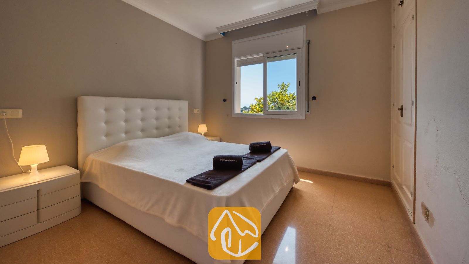 Vakantievilla's Costa Brava Spanje - Villa Valentina - Slaapkamer met een bed met een wit gecapitonneerd hoofdbord, twee nachtkastjes met kleine lampen en een raam dat uitzicht biedt op groen loof tegen een blauwe lucht.