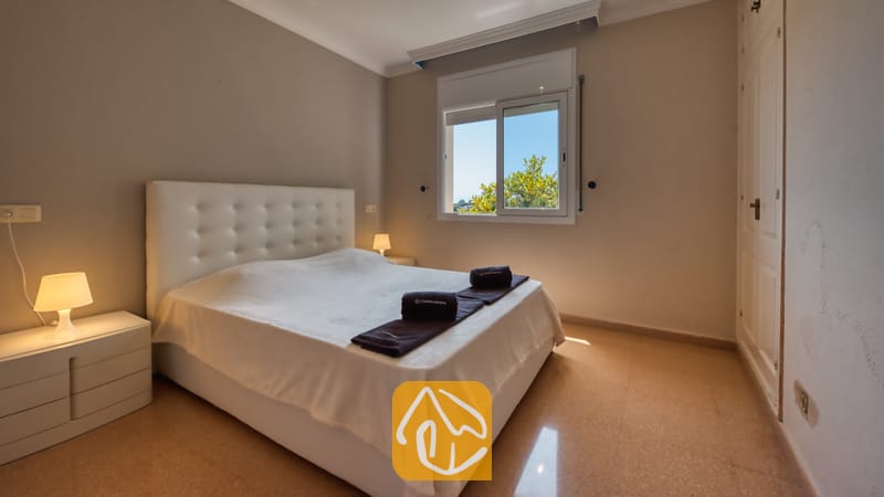 Villas de vacances Costa Brava Espagne - Villa Valentina - Chambre avec un lit doté d'une tête de lit capitonnée blanche, de deux tables de chevet surmontées de petites lampes, et d'une fenêtre encadrant un feuillage verdoyant sur fond de ciel bleu.