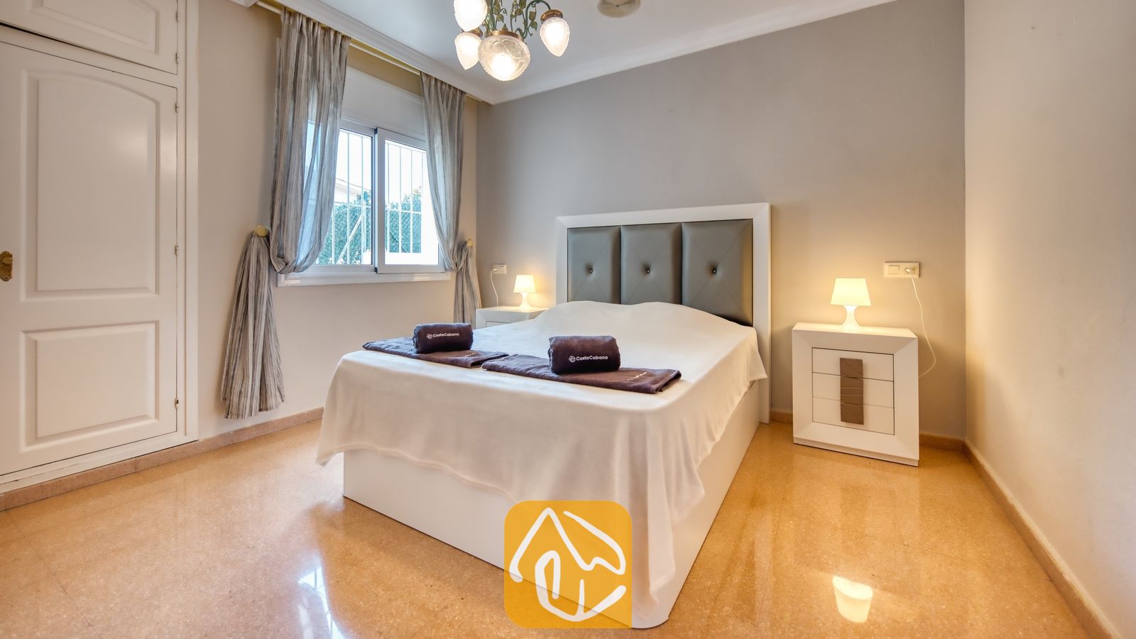 Vakantievilla's Costa Brava Spanje - Villa Valentina - Slaapkamer met een wit bed, een grijs gestoffeerd hoofdeinde, twee bijpassende nachtkastjes met lampen en een tegelvloer.