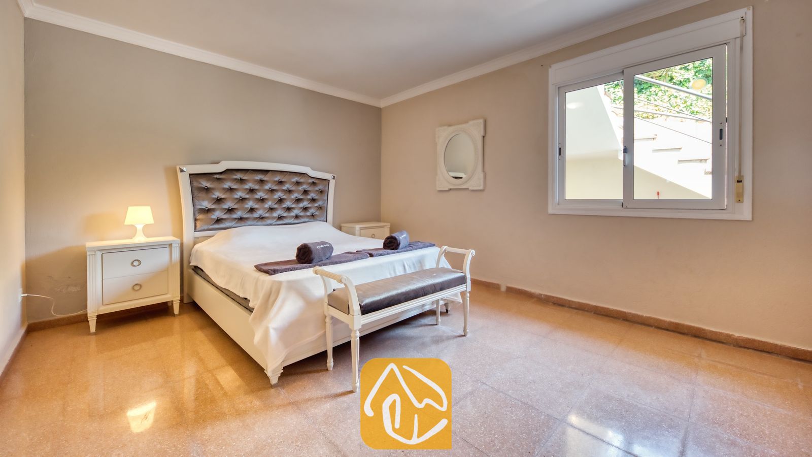 Vakantievilla's Costa Brava Spanje - Villa Valentina - Slaapkamer met een wit tweepersoonsbed met een gecapitonneerd grijs hoofdbord, een bijpassend nachtkastje met een verlichte lamp en een raam met uitzicht op een buitentrap.