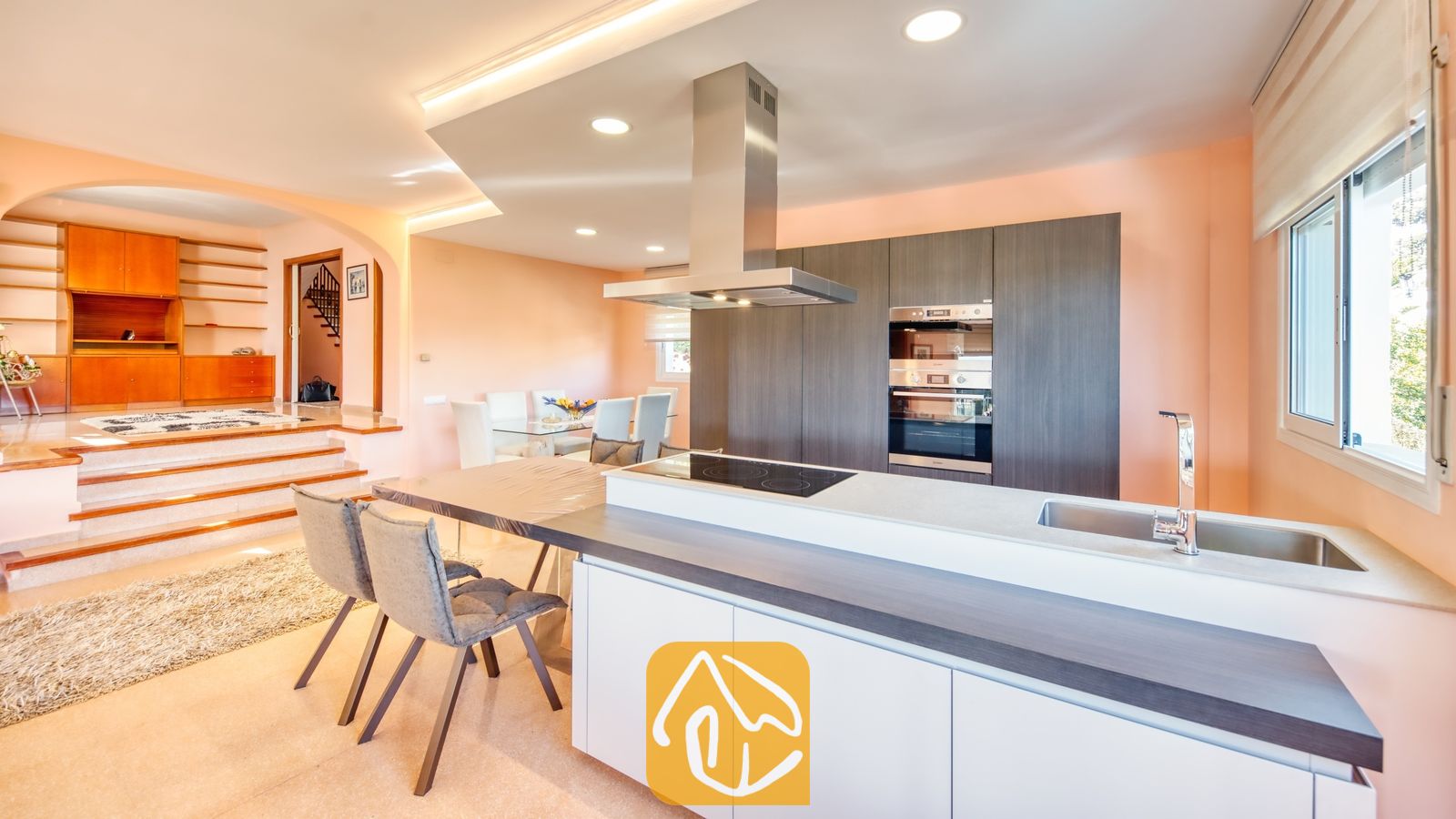 Vakantievilla's Costa Brava Spanje - Villa Valentina - Moderne keuken met donkere houten kasten, een roestvrijstalen afzuigkap en een aangrenzende eethoek met een trap naar een verhoogde woonkamer.