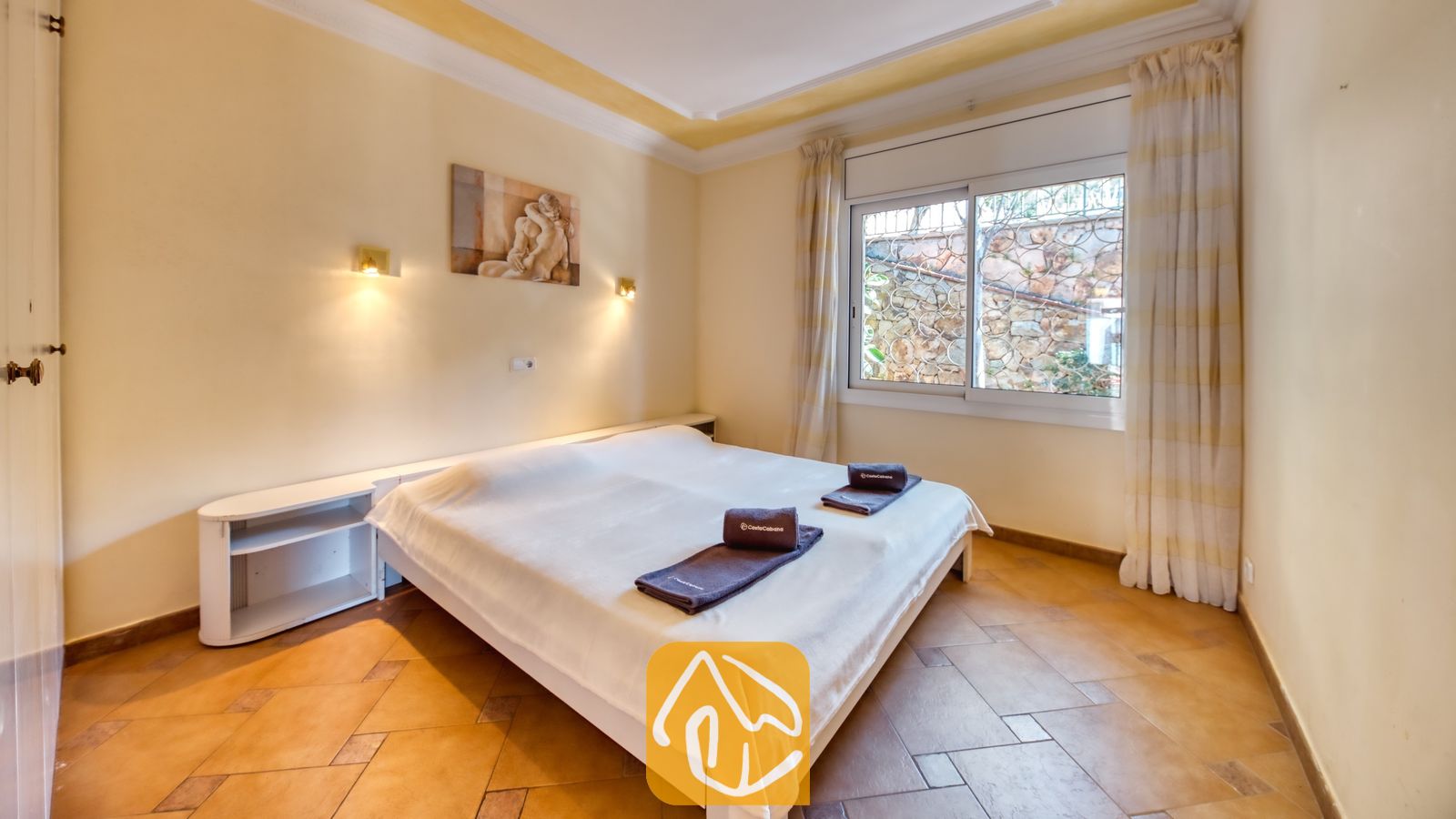 Vakantievilla's Costa Brava Spanje - Villa Donna - Slaapkamer met een tweepersoonsbed opgemaakt met witte lakens, donkere opgevouwen handdoeken, tegelvloeren en een raam met gordijnen.