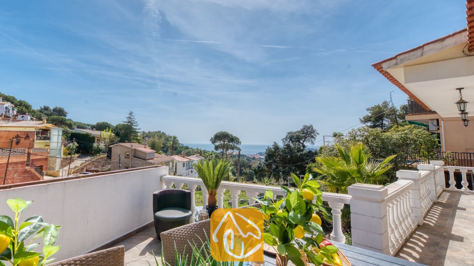 Vakantievilla's Costa Brava Spanje - Villa Donna - Balkonterras van Villa Donna met uitzicht over groene heuvels en de blauwe zee, voorzien van een eettafel met een in een pot geplante citroenboom en geweven stoelen onder een heldere hemel.