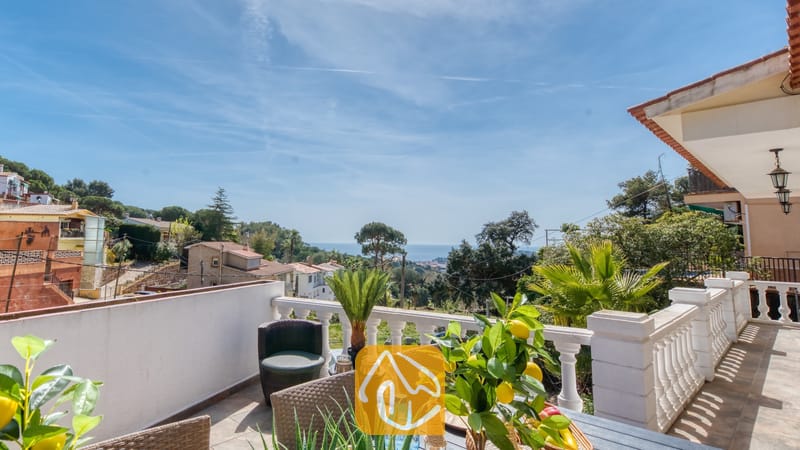 Vakantievilla's Costa Brava Spanje - Villa Donna - Balkonterras van Villa Donna met uitzicht over groene heuvels en de blauwe zee, voorzien van een eettafel met een in een pot geplante citroenboom en geweven stoelen onder een heldere hemel.