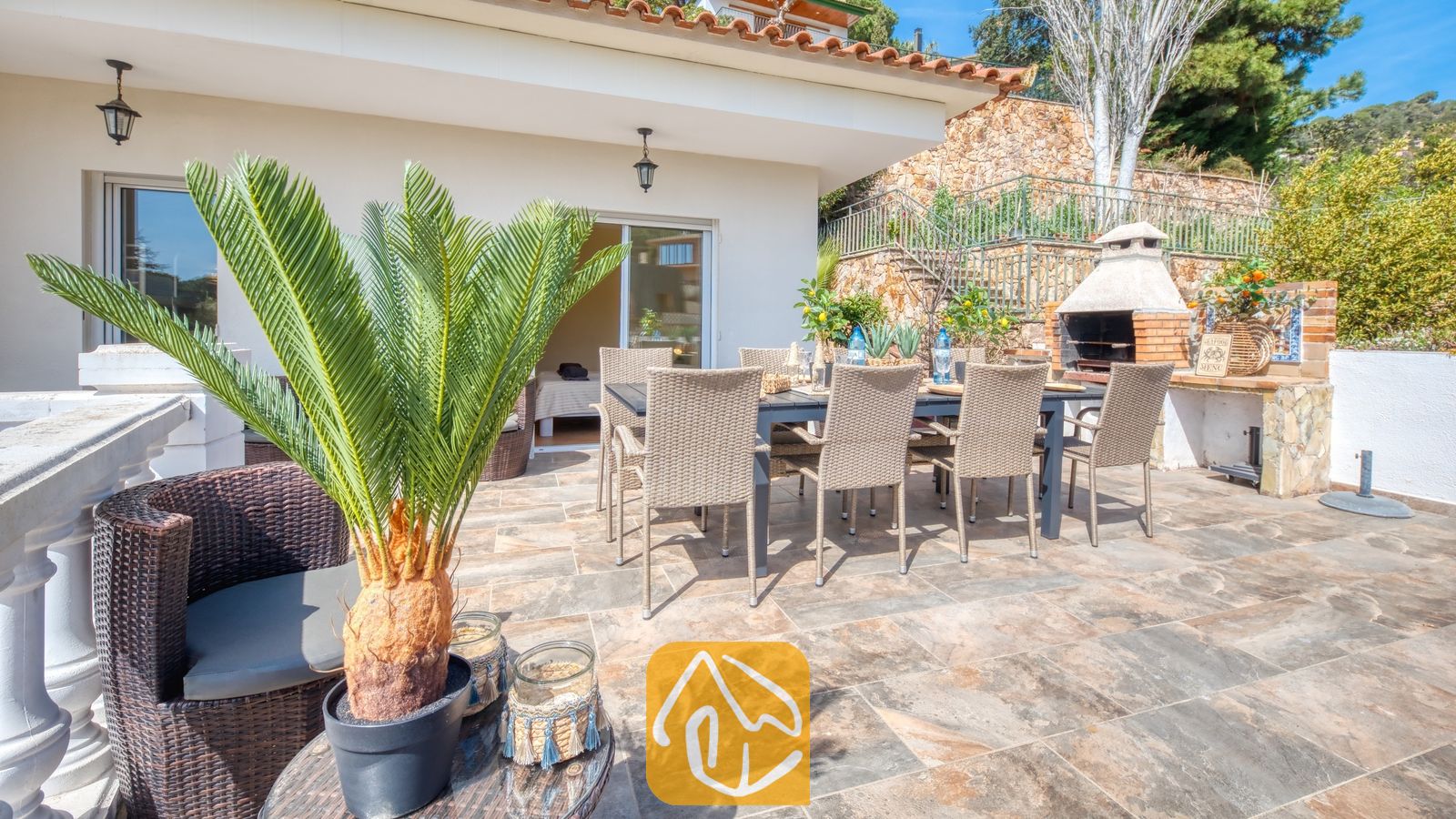 Vakantievilla's Costa Brava Spanje - Villa Donna - Zonnig terras bij Villa Donna, voorzien van een eettafel voor buiten met rieten stoelen, een ingebouwde stenen barbecue en betegelde vloeren.