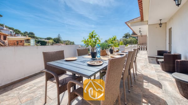 Vakantievilla's Costa Brava Spanje - Villa Donna - Terras van Villa Donna met een donkere eettafel en geweven rotan stoelen, gedekt met borden en potten met citroenboompjes onder een blauwe hemel.