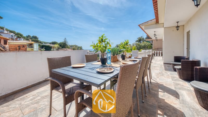 Vakantievilla's Costa Brava Spanje - Villa Donna - Terras van Villa Donna met een donkere eettafel en geweven rotan stoelen, gedekt met borden en potten met citroenboompjes onder een blauwe hemel.