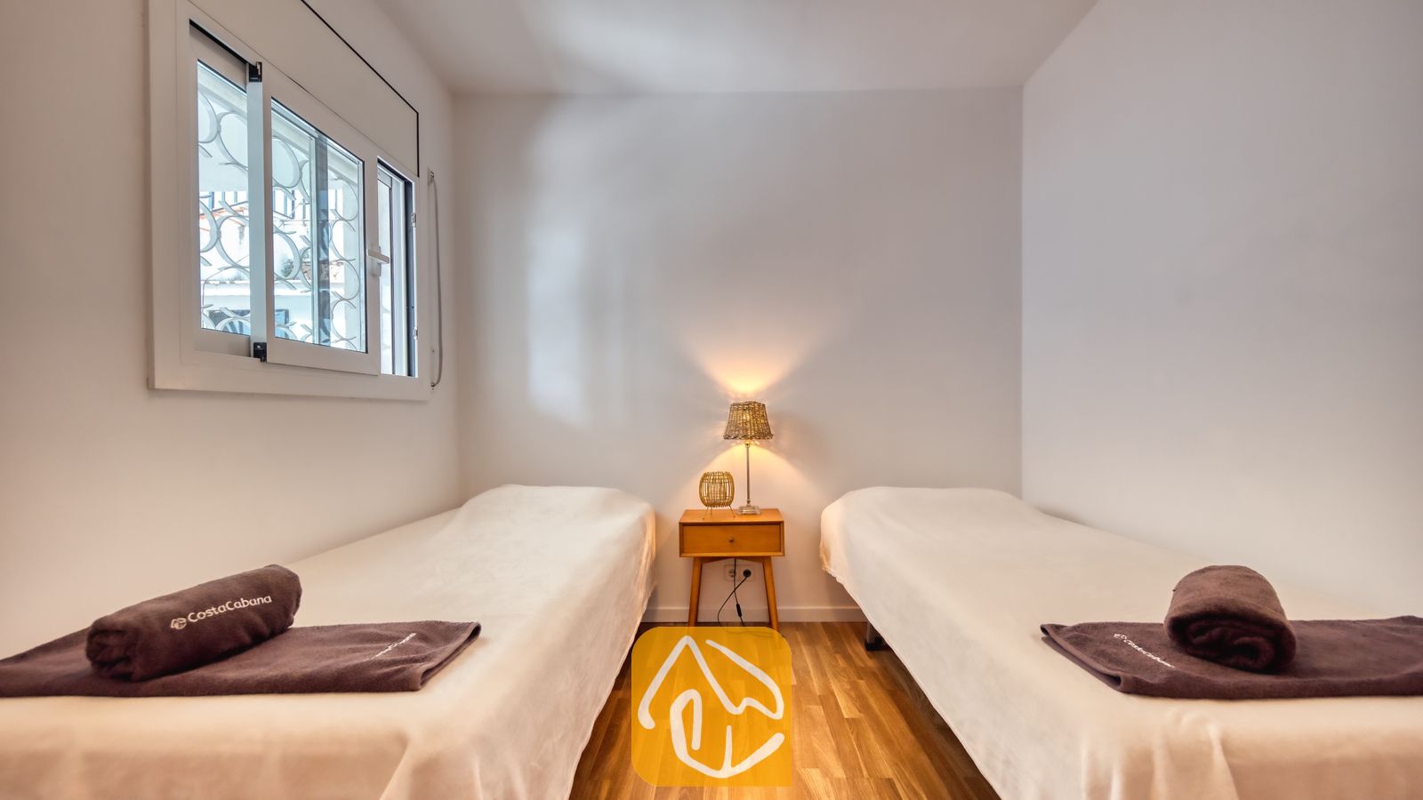 Vakantievilla's Costa Brava Spanje - Villa Donna - Slaapkamer met twee eenpersoonsbedden, voorzien van wit beddengoed, donkerbruine opgerolde handdoeken, een houten nachtkastje en een klein tafellampje.