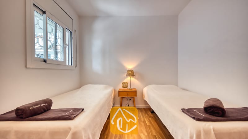 Holiday villas Costa Brava Spain - Villa Donna - Bedroom with two twin beds featuring white linens, dark brown rolled towels, a wooden bedside table, and a small table lamp.
