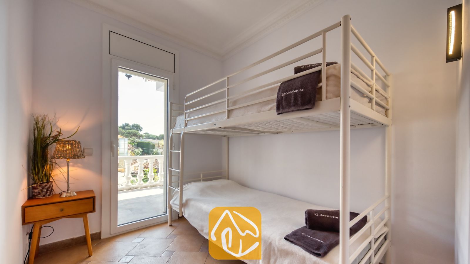 Vakantievilla's Costa Brava Spanje - Villa Donna - Slaapkamer in Villa Donna met een wit metalen stapelbed voorzien van opgevouwen donkere handdoeken, een houten nachtkastje met een gevlochten lamp en een glazen deur die uitkomt op een balkon.