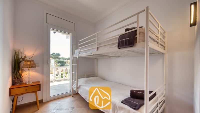 Holiday villas Costa Brava Spain - Villa Donna - Bedroom at Villa Donna featuring a white metal bunk bed with folded dark towels, a wooden side table with a woven lamp, and a glass door opening to a balcony.