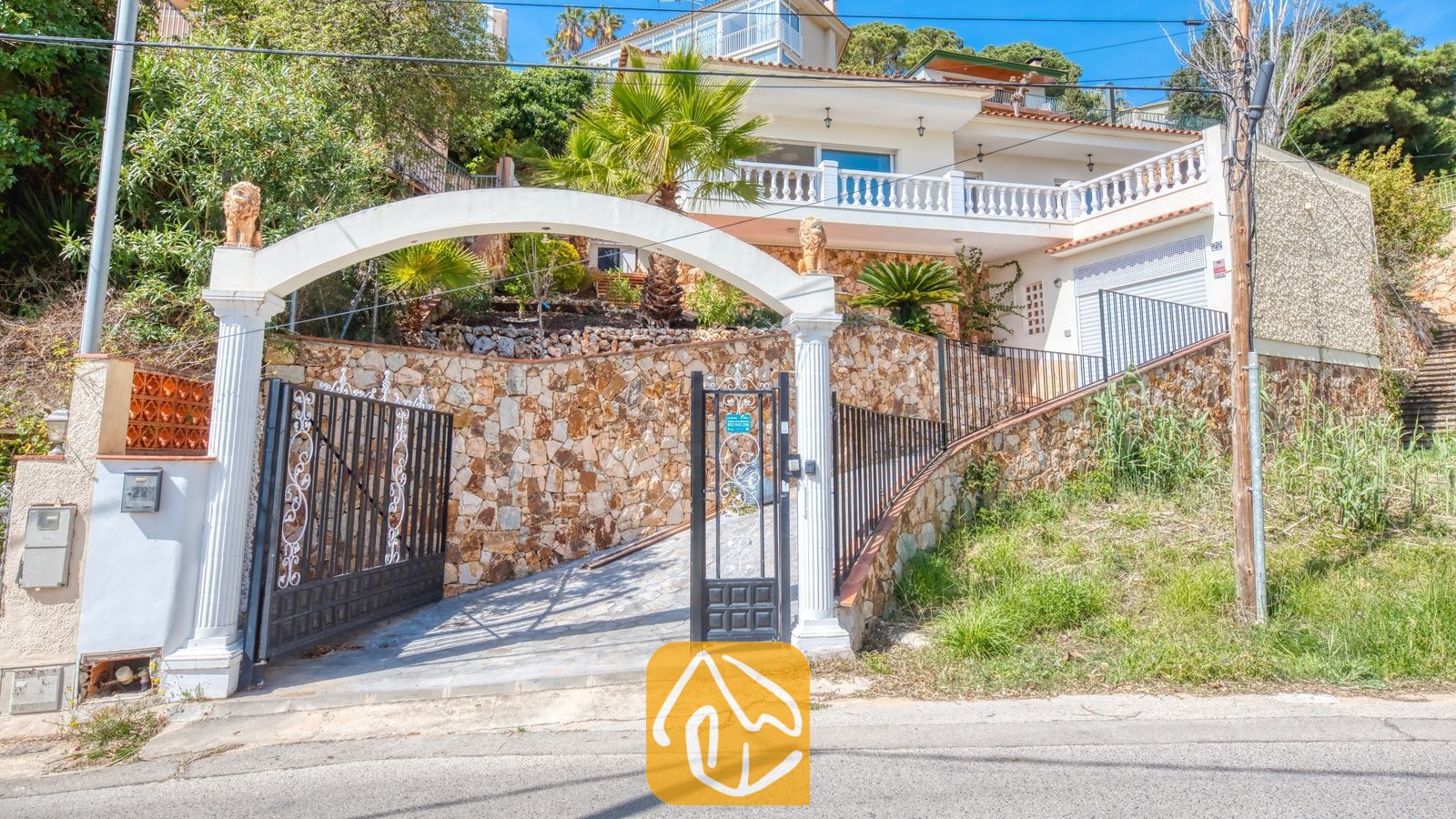 Vakantievilla's Costa Brava Spanje - Villa Donna - Buitenkant van Villa Donna met een witte boogvormige ingang, zwart metalen hekken, stenen muren en een villa met meerdere verdiepingen en een balkon tegen een heuvel.