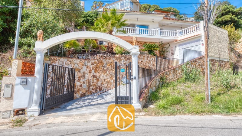 Holiday villas Costa Brava Spain - Villa Donna - Exterior entrance of Villa Donna featuring a white arched gateway, black metal gates, stone walls, and a multi-level villa with a balcony set against a hillside.