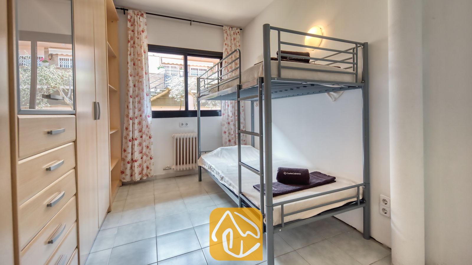 Vakantievilla's Costa Brava Spanje - Casa Costa - Slaapkamer, ingericht met een grijs metalen stapelbed, een lichte houten kledingkast met lades en bloemendecoratie gordijnen die het raam omlijsten.