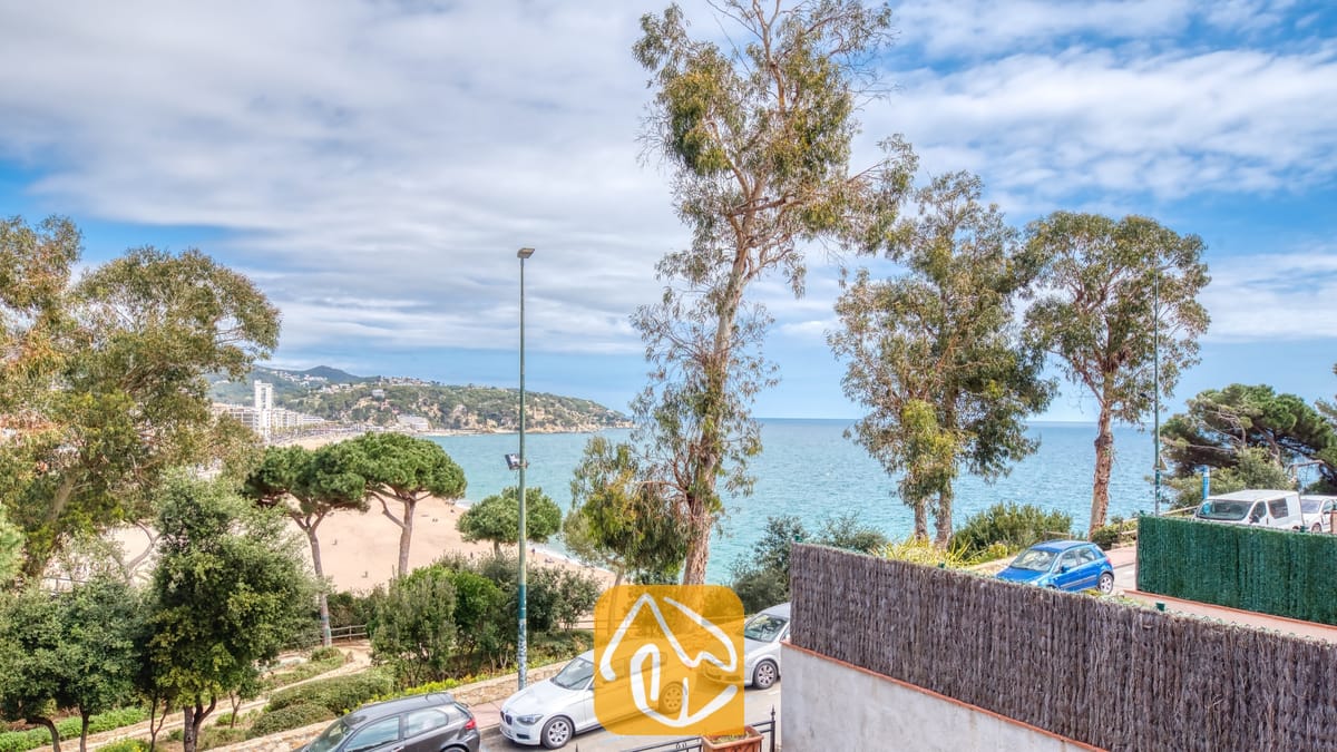 Vakantievilla's Costa Brava Spanje - Casa Costa - Uitzicht vanuit Casa Costa met hoge bomen, een zandstrand en de blauwe zee onder een licht bewolkte hemel.