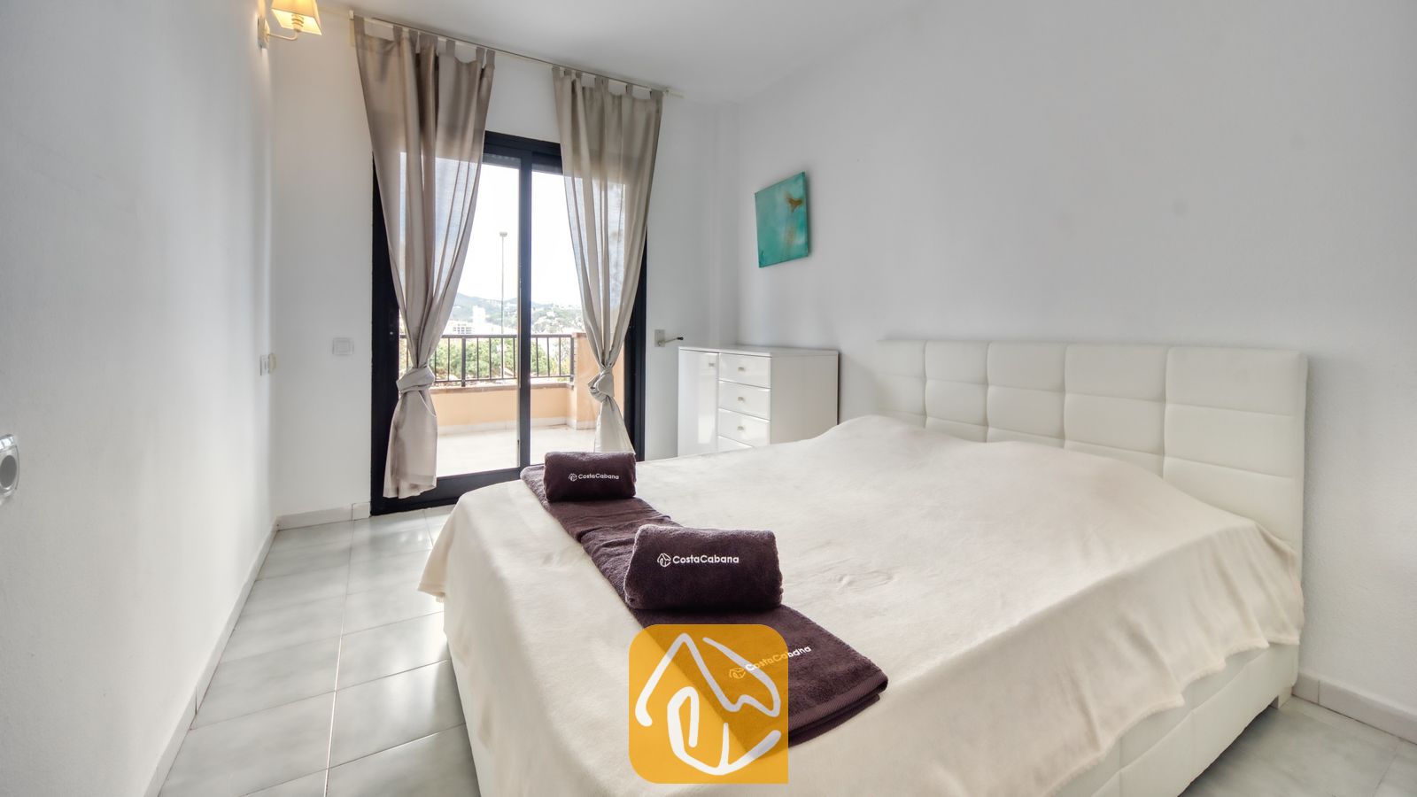 Vakantievilla's Costa Brava Spanje - Casa Costa - Slaapkamer met een wit gestoffeerd hoofdbord, opgerolde bruine handdoeken op het bed en een glazen deur die uitkomt op een balkon.