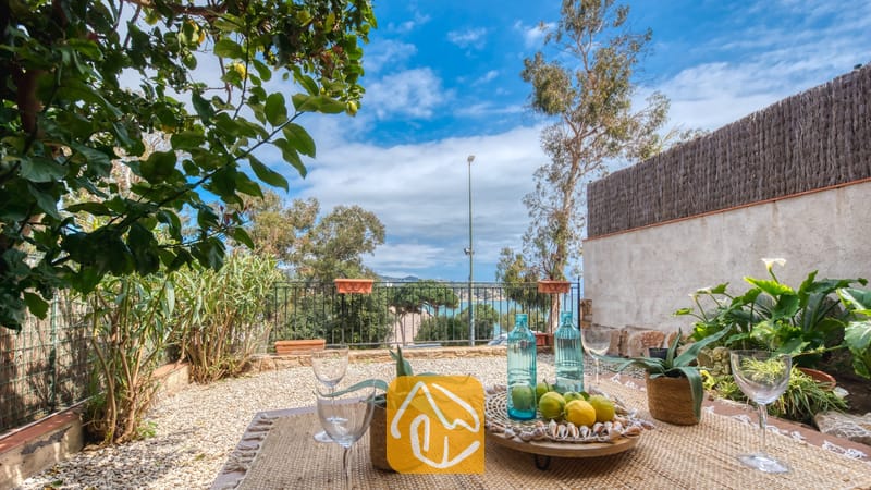 Vakantievilla's Costa Brava Spanje - Casa Costa - Terras van Casa Costa met een tafel gedecoreerd met wijnglazen, blauwe glazen flessen en een dienblad met citroenen, uitkijkend op bomen en de zee onder een blauwe lucht.