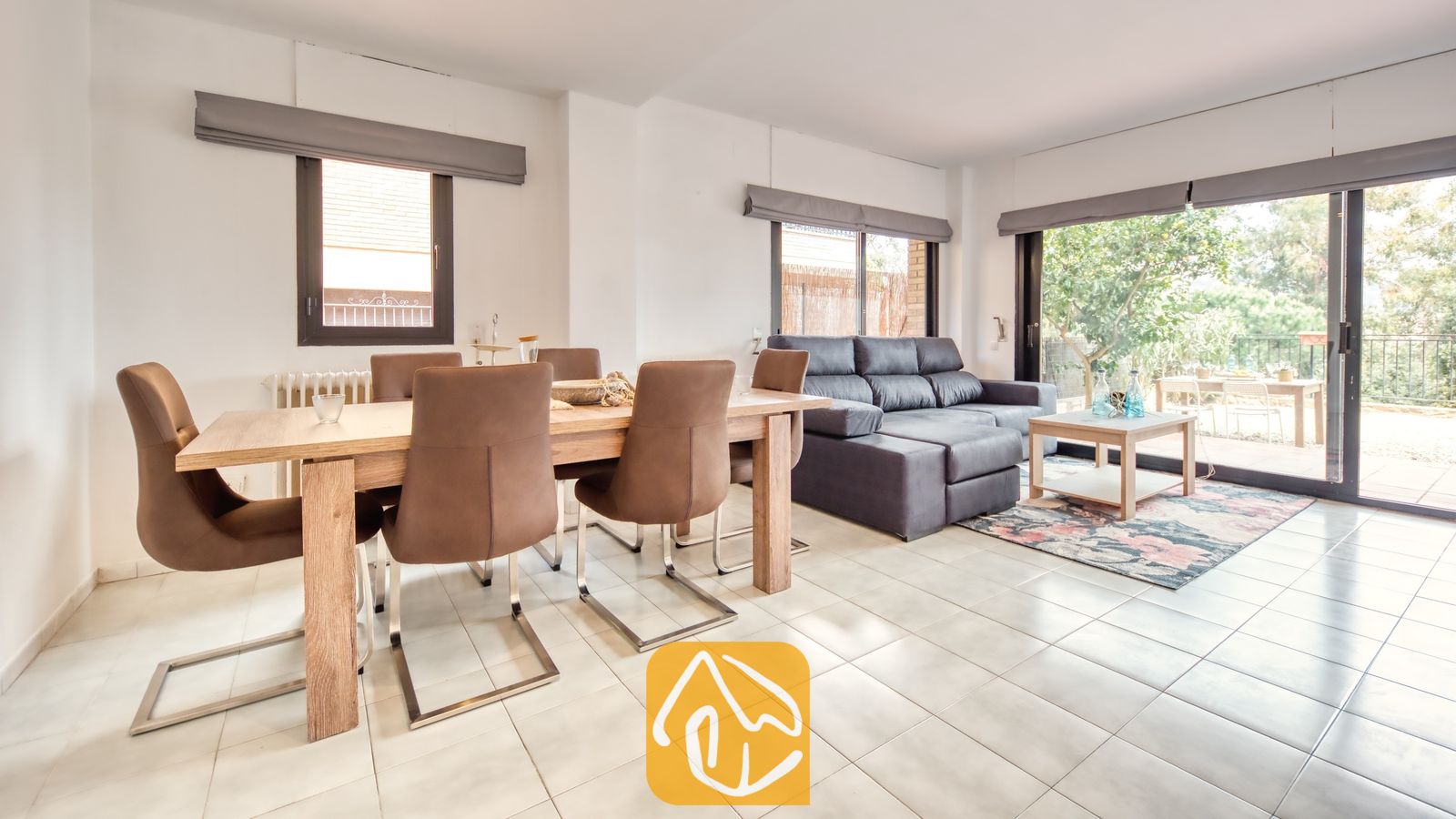 Vakantievilla's Costa Brava Spanje - Casa Costa - Open woon- en eetgedeelte bij Casa Costa, voorzien van een houten eettafel met bruine stoelen en een grijze bank bij de glazen schuifdeuren.