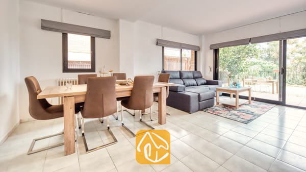 Vakantievilla's Costa Brava Spanje - Casa Costa - Open woon- en eetgedeelte bij Casa Costa, voorzien van een houten eettafel met bruine stoelen en een grijze bank bij de glazen schuifdeuren.