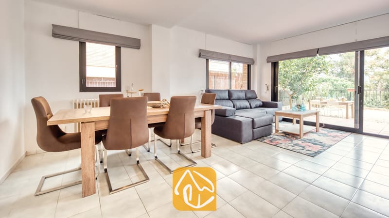 Vakantievilla's Costa Brava Spanje - Casa Costa - Open woon- en eetgedeelte bij Casa Costa, voorzien van een houten eettafel met bruine stoelen en een grijze bank bij de glazen schuifdeuren.