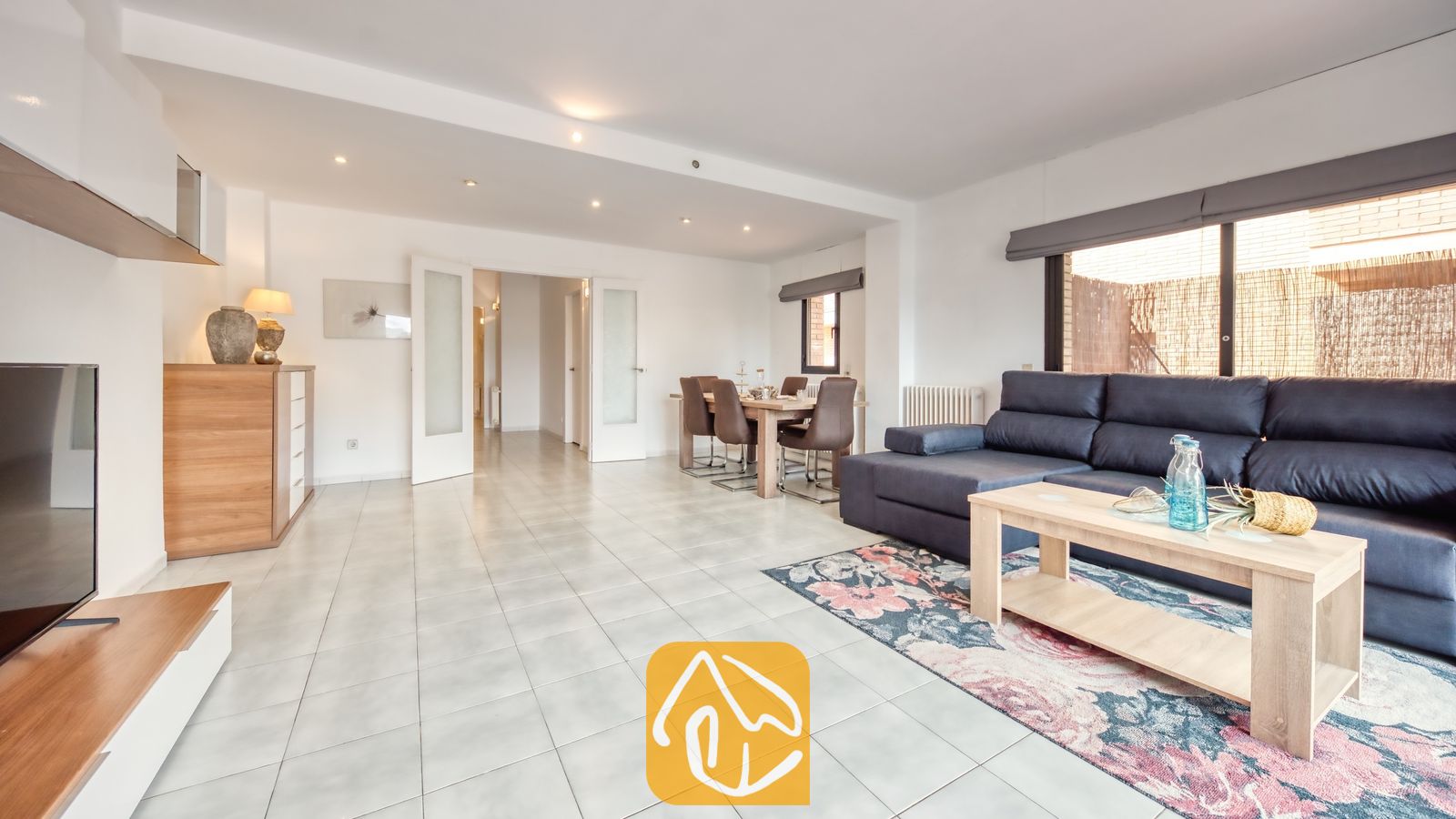 Vakantievilla's Costa Brava Spanje - Casa Costa - Ruime woon- en eetkamer bij Casa Costa met een donkerblauwe hoekbank, een lichte houten salontafel en een bloemetjestapijt op een tegelvloer.