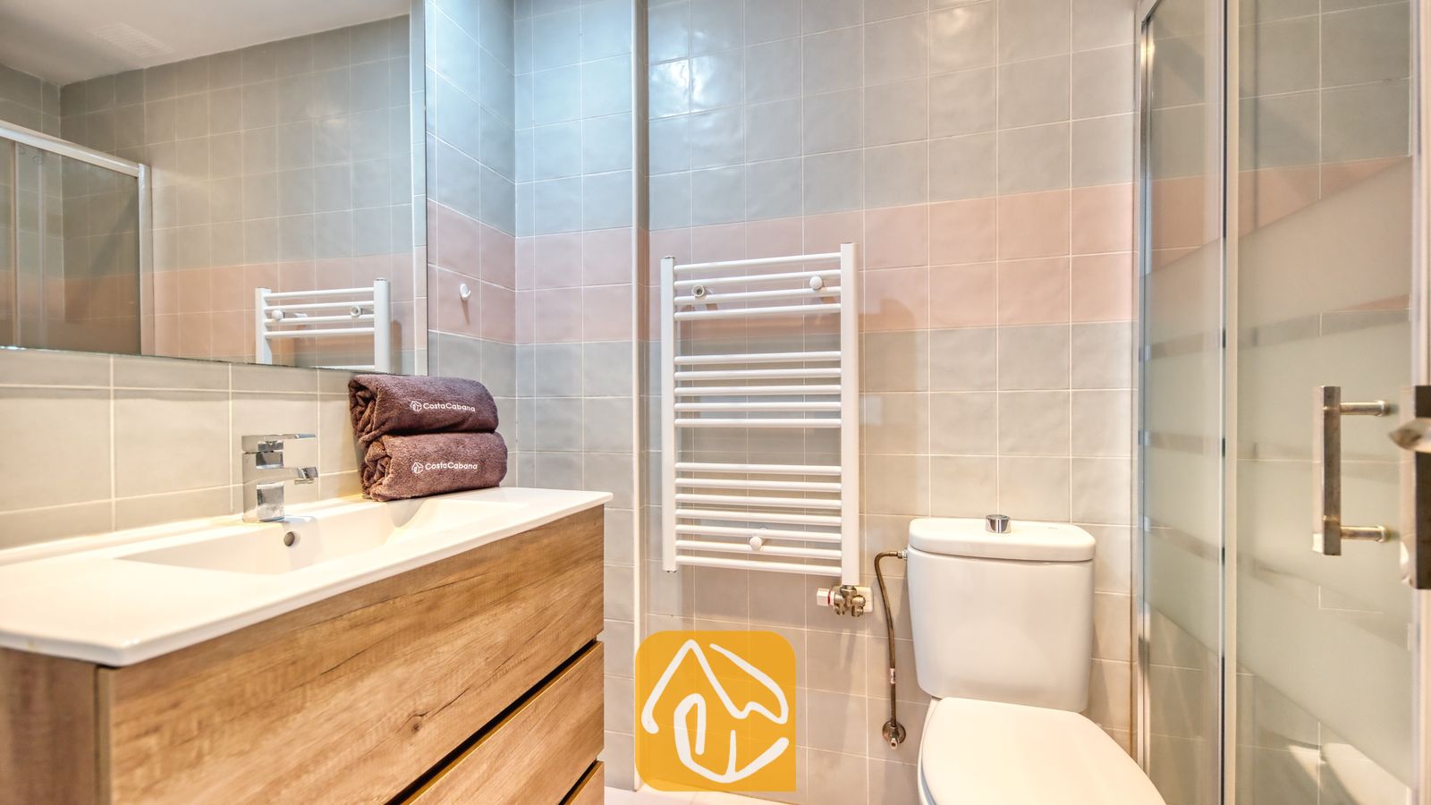 Vakantievilla's Costa Brava Spanje - Casa Costa - Moderne badkamer met een houten badkamermeubel en een witte wastafel, een wandgemonteerd wit handdoekenrek en betegelde muren in grijze en roze tinten.
