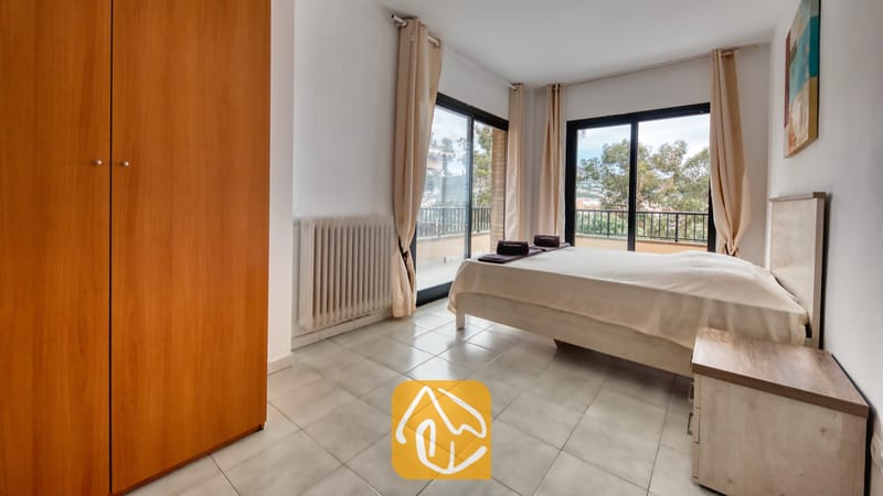 Vakantievilla's Costa Brava Spanje - Casa Costa - Slaapkamer met tegelvloer, een tweepersoonsbed, een grote houten kledingkast en glazen schuifdeuren die open kunnen naar een balkon met uitzicht op bomen.