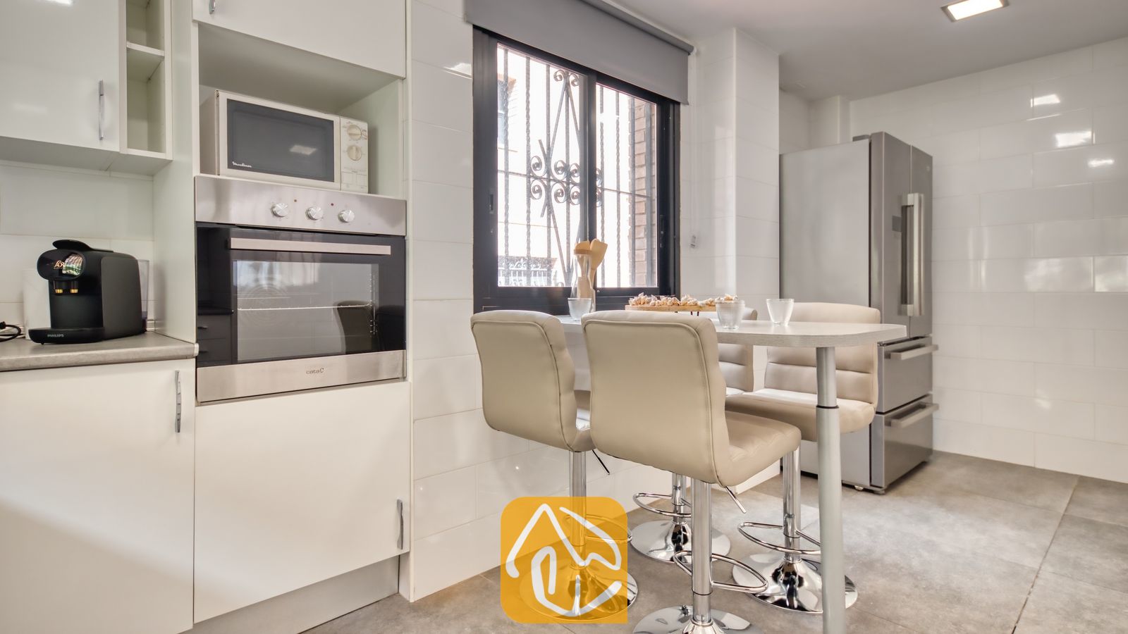 Vakantievilla's Costa Brava Spanje - Casa Costa - Moderne keuken met witte betegelde muren, een inbouw oven van roestvrij staal, een magnetron en een hoge eettafel met vier beige barstoelen.