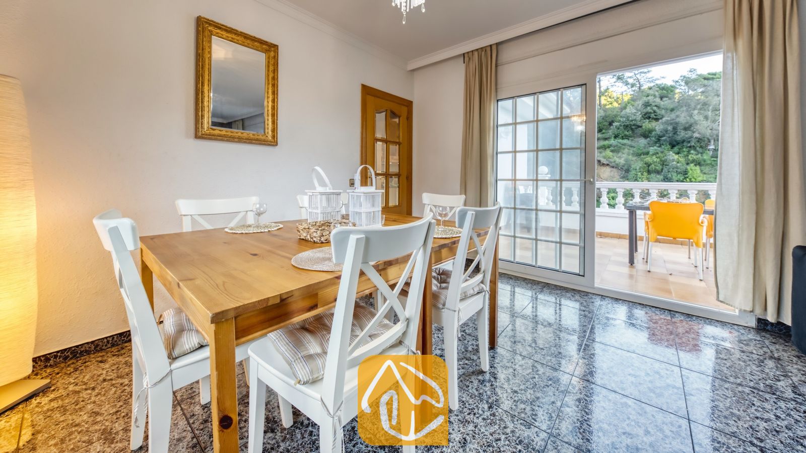 Vakantievilla's Costa Brava Spanje - Villa Rosa - Houten eettafel, omringd door witte stoelen met kussens, opgesteld in een lichte eethoek binnen in Villa Rosa, met glazen schuifdeuren die uitkomen op een terras.