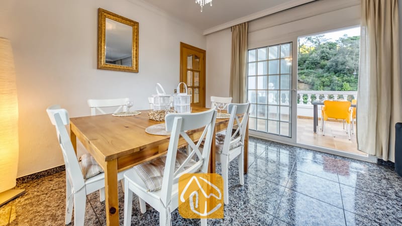 Vakantievilla's Costa Brava Spanje - Villa Rosa - Houten eettafel, omringd door witte stoelen met kussens, opgesteld in een lichte eethoek binnen in Villa Rosa, met glazen schuifdeuren die uitkomen op een terras.