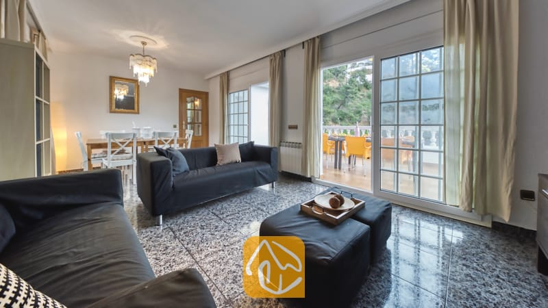 Vakantievilla's Costa Brava Spanje - Villa Rosa - Ruime woonkamer in Villa Rosa met donkergrijze banken, gepolijste stenen tegelvloeren en grote glazen schuifdeuren die uitkomen op een buitenterras.
