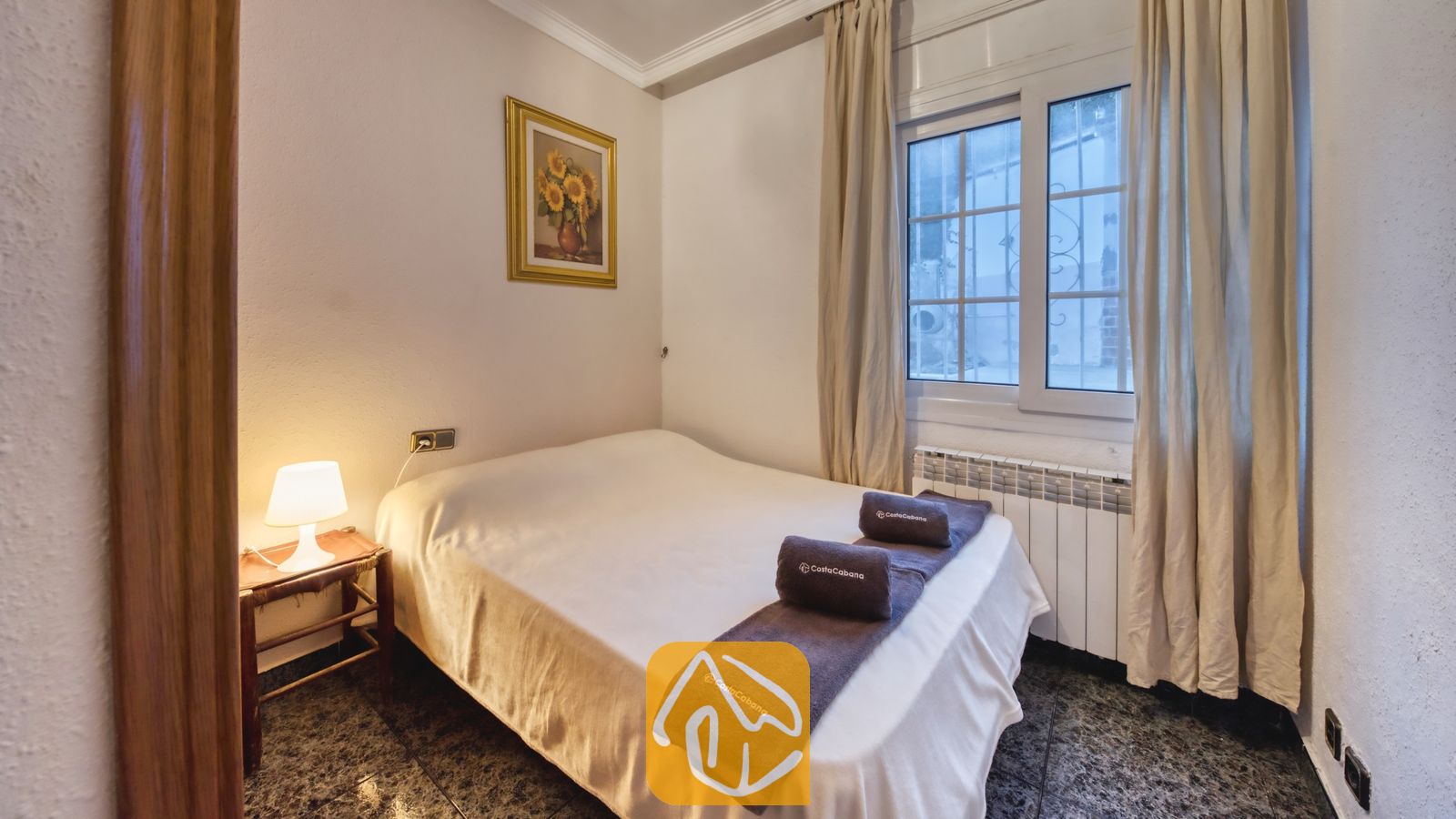 Vakantievilla's Costa Brava Spanje - Villa Rosa - Slaapkamer met een tweepersoonsbed opgemaakt met witte lakens, donker opgevouwen handdoeken, een nachtkastje met een klein lampje en een ingelijst zonnebloemschilderij.