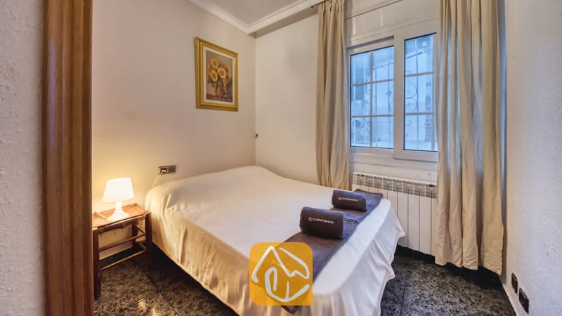 Vakantievilla's Costa Brava Spanje - Villa Rosa - Slaapkamer met een tweepersoonsbed opgemaakt met witte lakens, donker opgevouwen handdoeken, een nachtkastje met een klein lampje en een ingelijst zonnebloemschilderij.