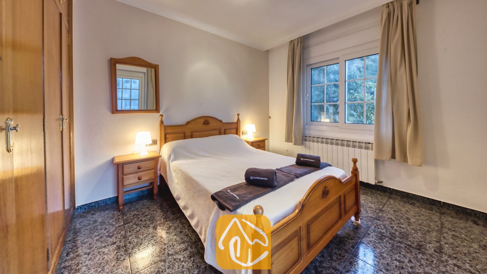 Vakantievilla's Costa Brava Spanje - Villa Rosa - Slaapkamer met een houten bedframe, twee kleine nachtkastjes met sfeervolle lampjes, opgerolde handdoeken op het witte dekbed, een wandspiegel en een raam met beige gordijnen.