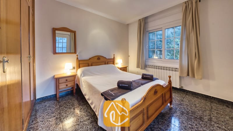 Vakantievilla's Costa Brava Spanje - Villa Rosa - Slaapkamer met een houten bedframe, twee kleine nachtkastjes met sfeervolle lampjes, opgerolde handdoeken op het witte dekbed, een wandspiegel en een raam met beige gordijnen.