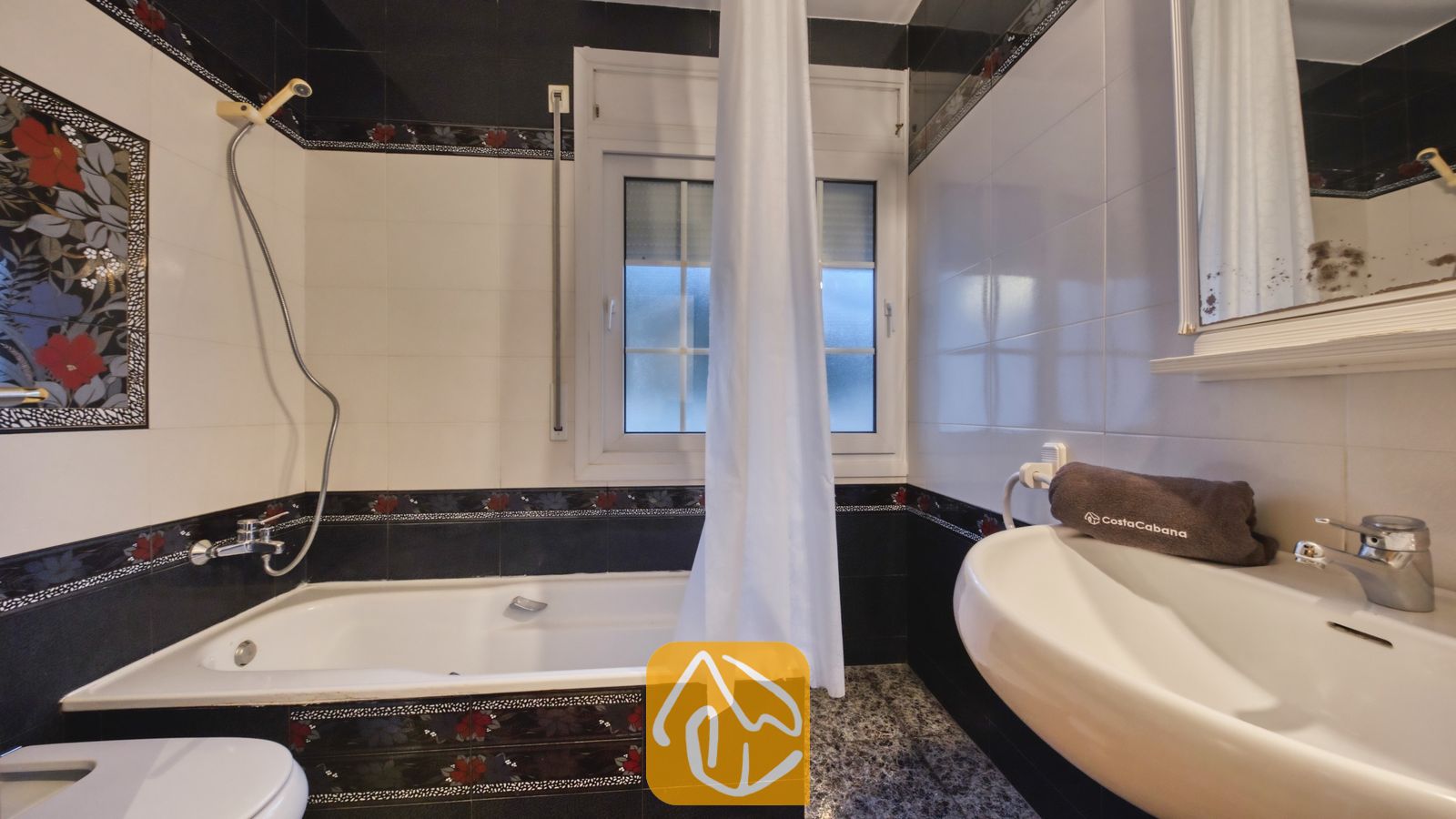 Vakantievilla's Costa Brava Spanje - Villa Rosa - Badkamer met een wit ligbad, aangesloten douchekop en zwart-wit betegelde muren, gedecoreerd met rode bloemmotieven.